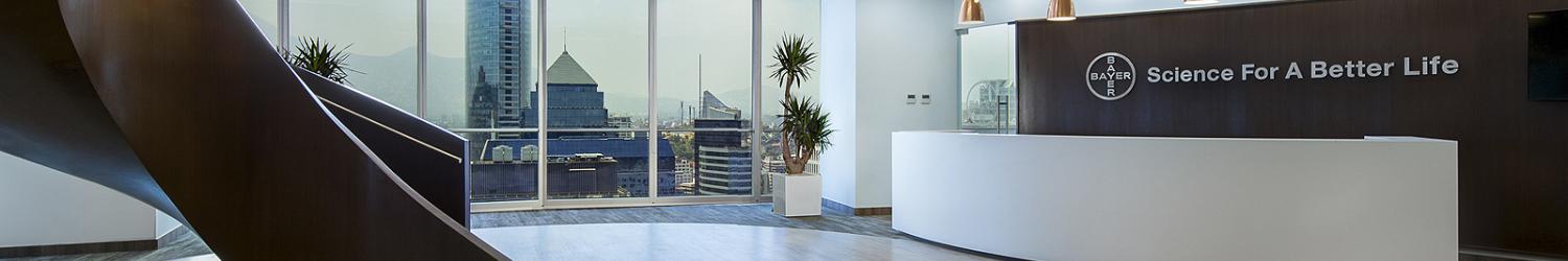 Moderno lobby de oficina con una gran escalera de caracol, plantas, un mostrador de recepción con el logotipo y el lema "Ciencia para una vida mejor" y vista de los rascacielos a través de ventanas de piso a techo.
