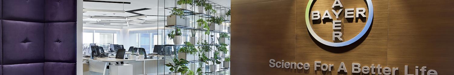 Interior de oficina moderno con mamparas de cristal y estanterías para plantas. El logotipo de Bayer y el eslogan "Ciencia para una vida mejor" se muestran en una pared revestida de madera.