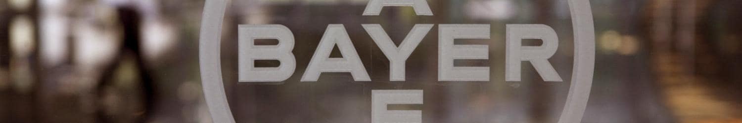 Primer plano del logotipo de Bayer sobre una superficie de vidrio, con una figura borrosa y un entorno visible en el fondo.