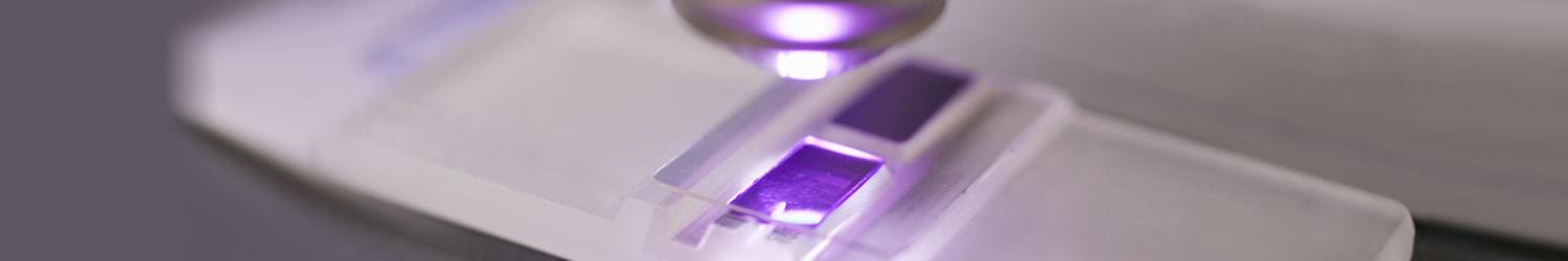 Primer plano de un portaobjetos de microscopio con una luz violeta que ilumina una muestra bajo una lente.