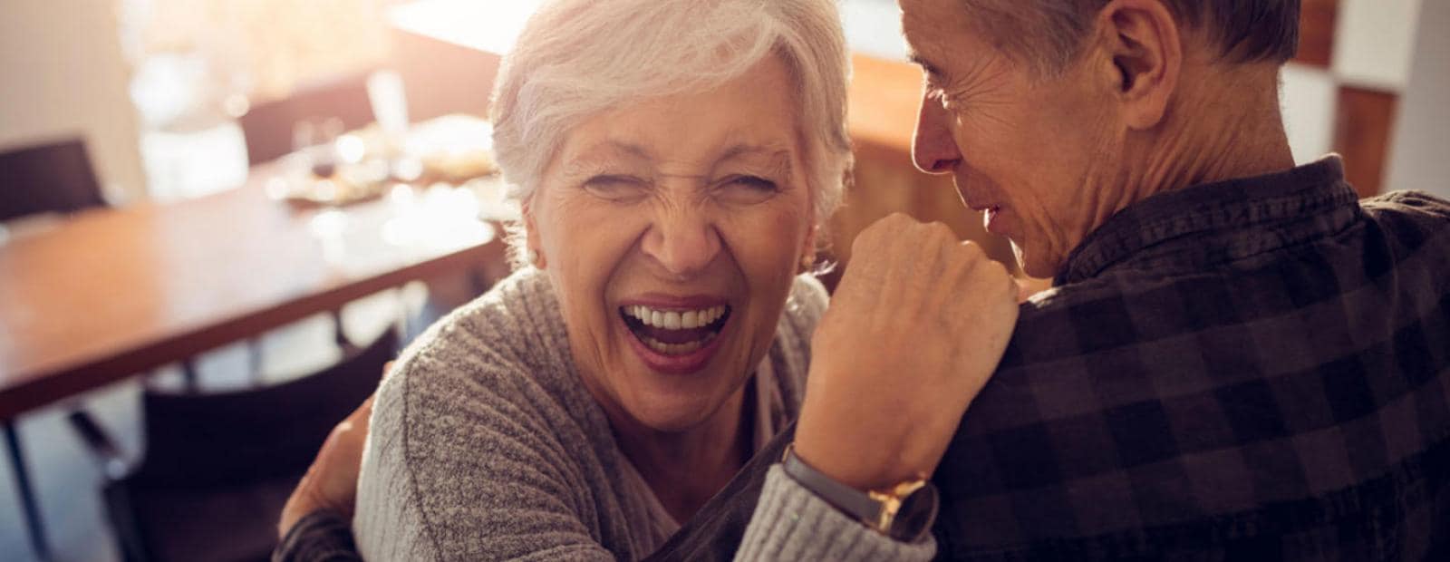Una anciana se ríe mientras abraza a un anciano en una habitación cálidamente iluminada.