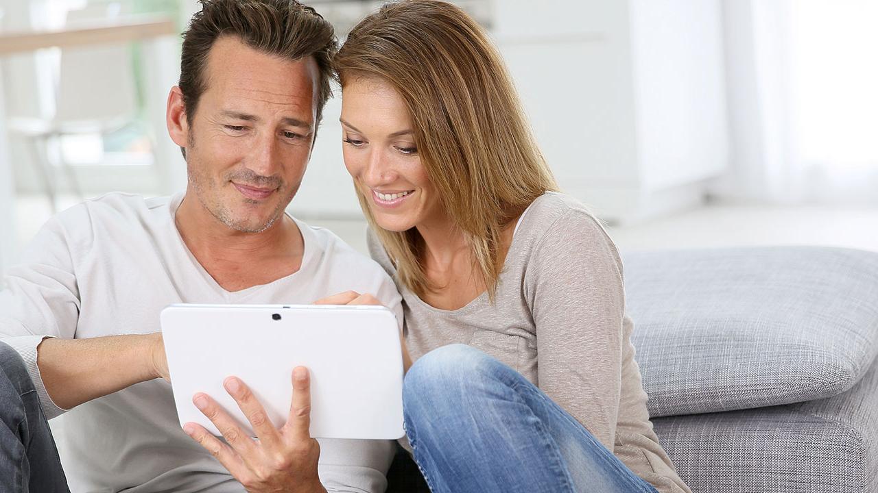 Una pareja se sienta en el suelo, sonriendo mientras miran juntos una tableta en una habitación luminosa.