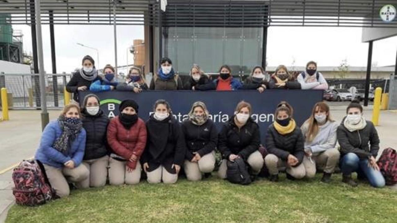 Un grupo de 17 personas con máscaras posan frente a un edificio, arrodillados y de pie en dos filas, con un gran cartel detrás de ellos.