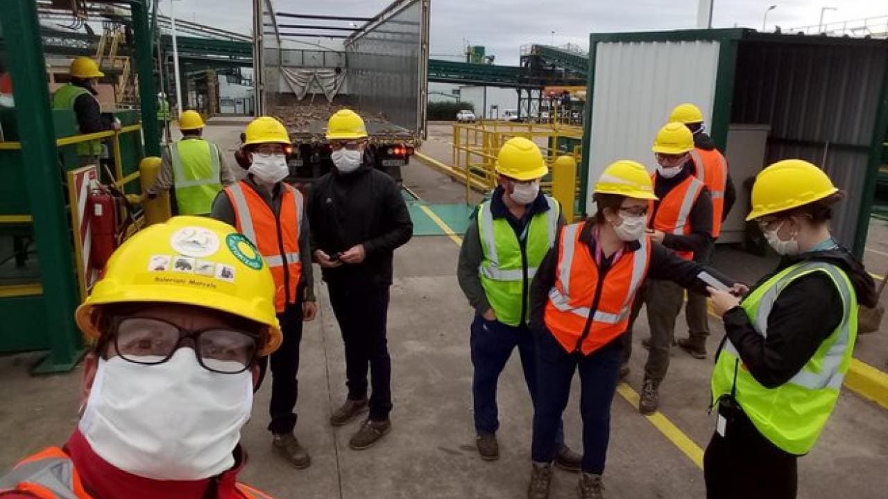 Un grupo de trabajadores que usan equipo de seguridad, incluidos cascos, máscaras y chalecos, se encuentran cerca de un sitio industrial con maquinaria y un camión al fondo.