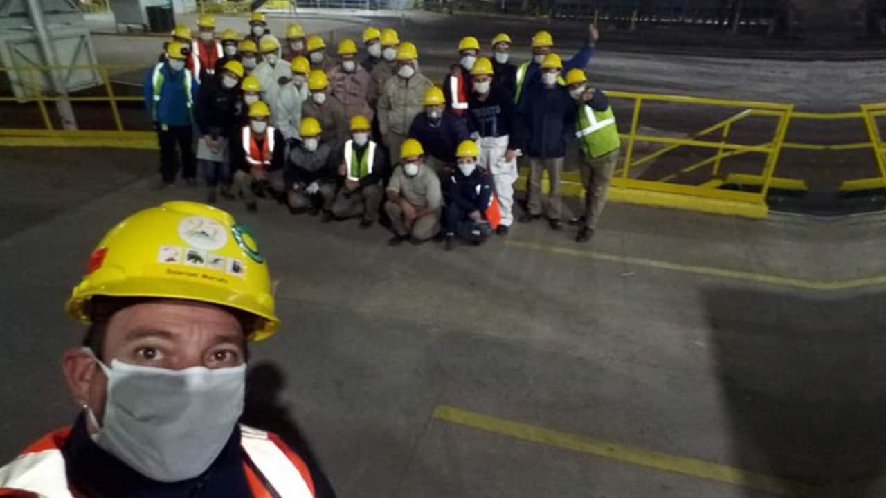 Un grupo de personas con cascos de construcción y máscaras faciales posan para una foto en un interior, con un hombre delante tomando la fotografía.