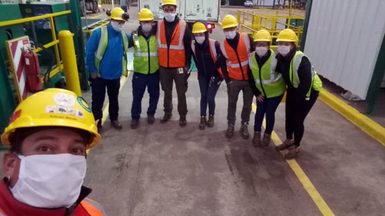 Un grupo de personas con cascos, máscaras y chalecos de seguridad posan para una foto en un entorno industrial. Una persona se toma un selfie en primer plano.