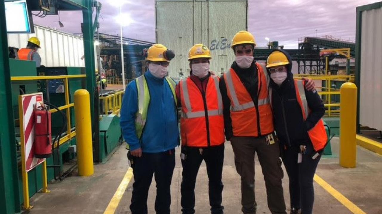 Cuatro trabajadores de la construcción con cascos y chalecos de seguridad se encuentran juntos en una zona industrial. Llevan mascarillas y el fondo muestra equipos industriales y un cielo nublado.