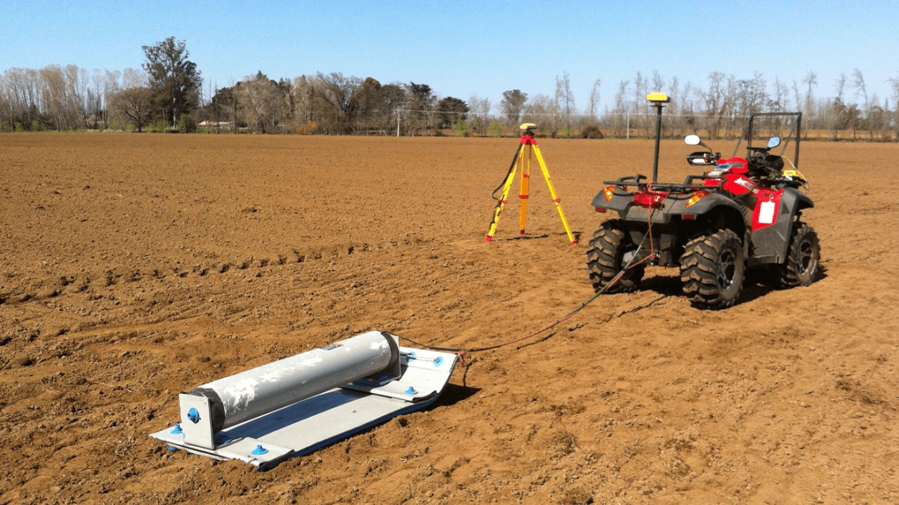 Un vehículo todo terreno arrastra un dispositivo de radar de penetración terrestre a lo largo de un campo grande y plano, con un trípode de reconocimiento cerca, bajo un cielo azul claro.