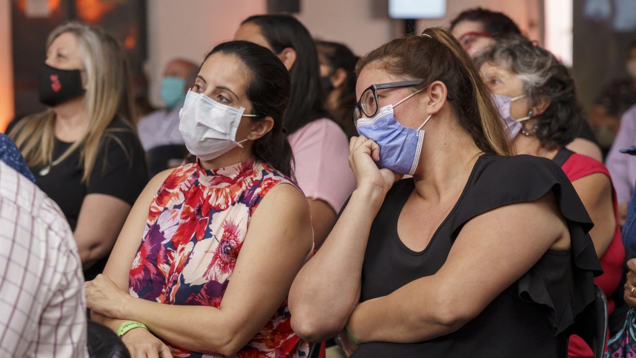 Miembros de la audiencia con máscaras sentados y observando atentamente un evento en el interior.