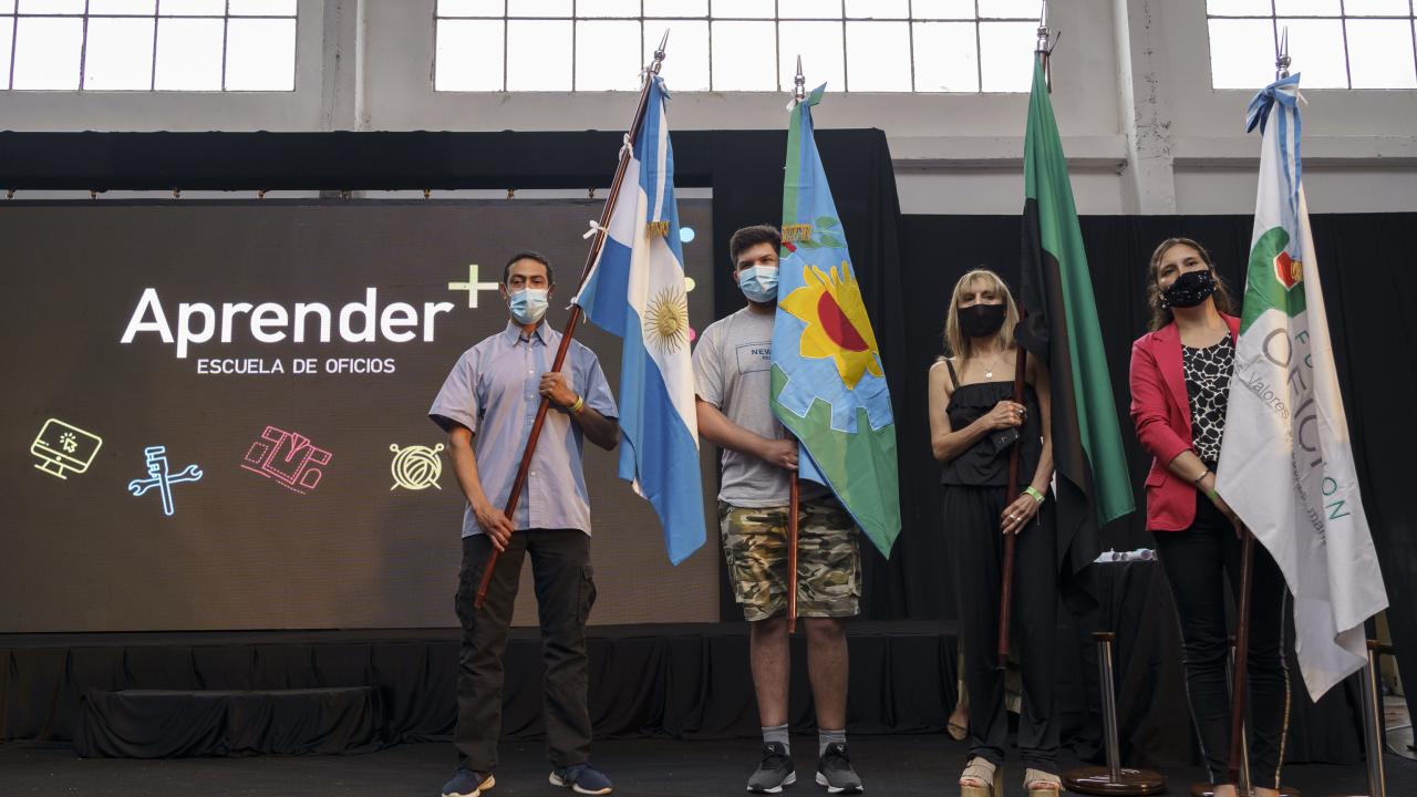 Cuatro personas están en el escenario sosteniendo banderas en un evento, con una pancarta que dice "Aprender+" en el fondo.