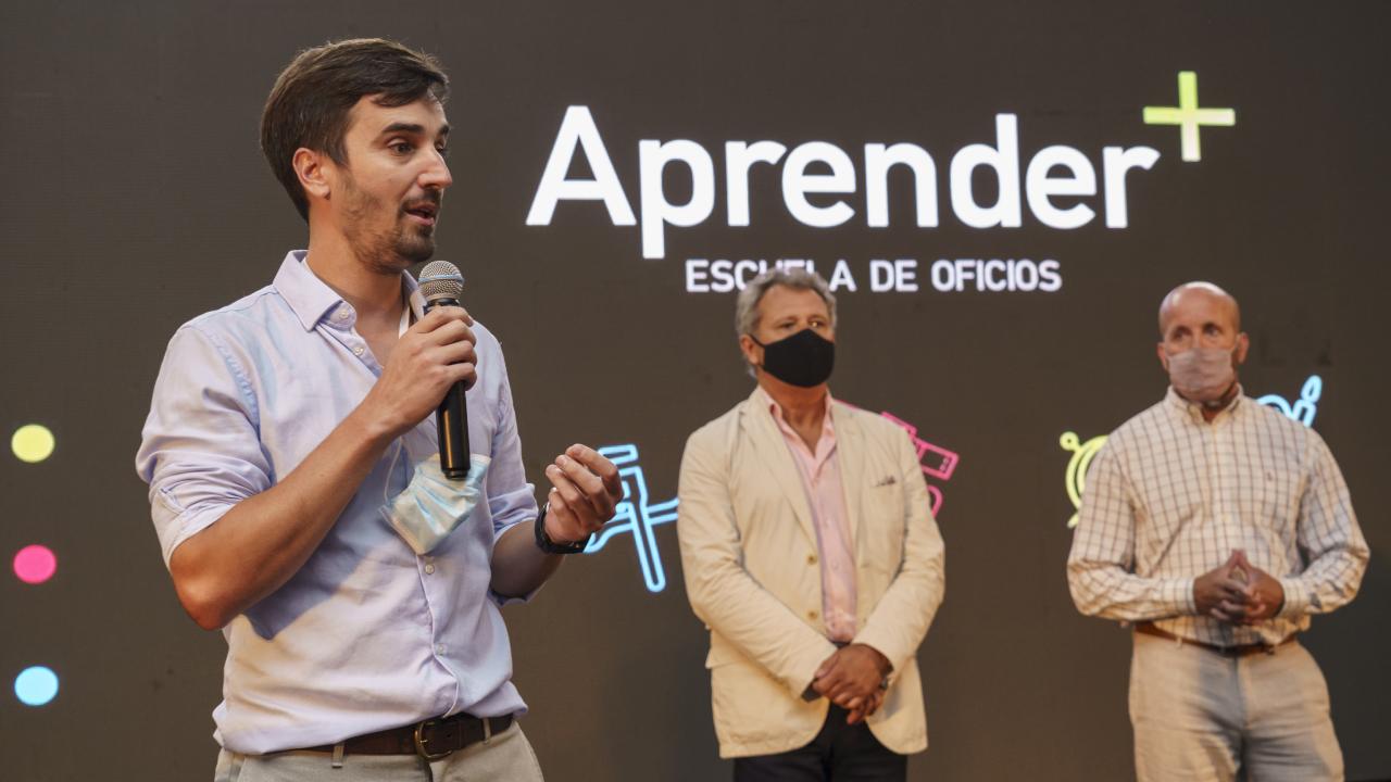Un hombre habla por un micrófono en el escenario junto a otros dos hombres. En el fondo, una pantalla muestra "Aprender Escuela de Oficios".