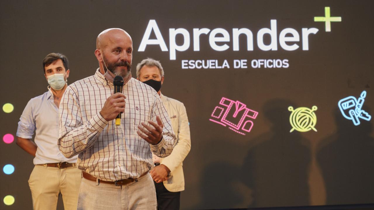 Un hombre habla por un micrófono en el escenario durante el evento Aprender Escuela de Oficios. Dos hombres están detrás de él; uno lleva una mascarilla.