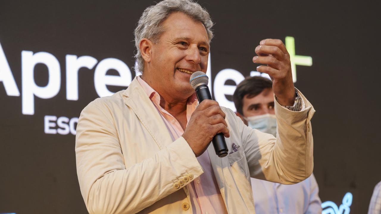 Un hombre con una chaqueta beige sostiene un micrófono y hace gestos con la otra mano mientras habla. Una persona en el fondo lleva una mascarilla. El texto de fondo dice "Aprende+".