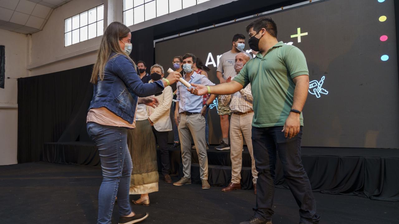 Una mujer entrega un certificado a un hombre en el escenario, con varias personas de pie en el fondo, todas con máscaras.