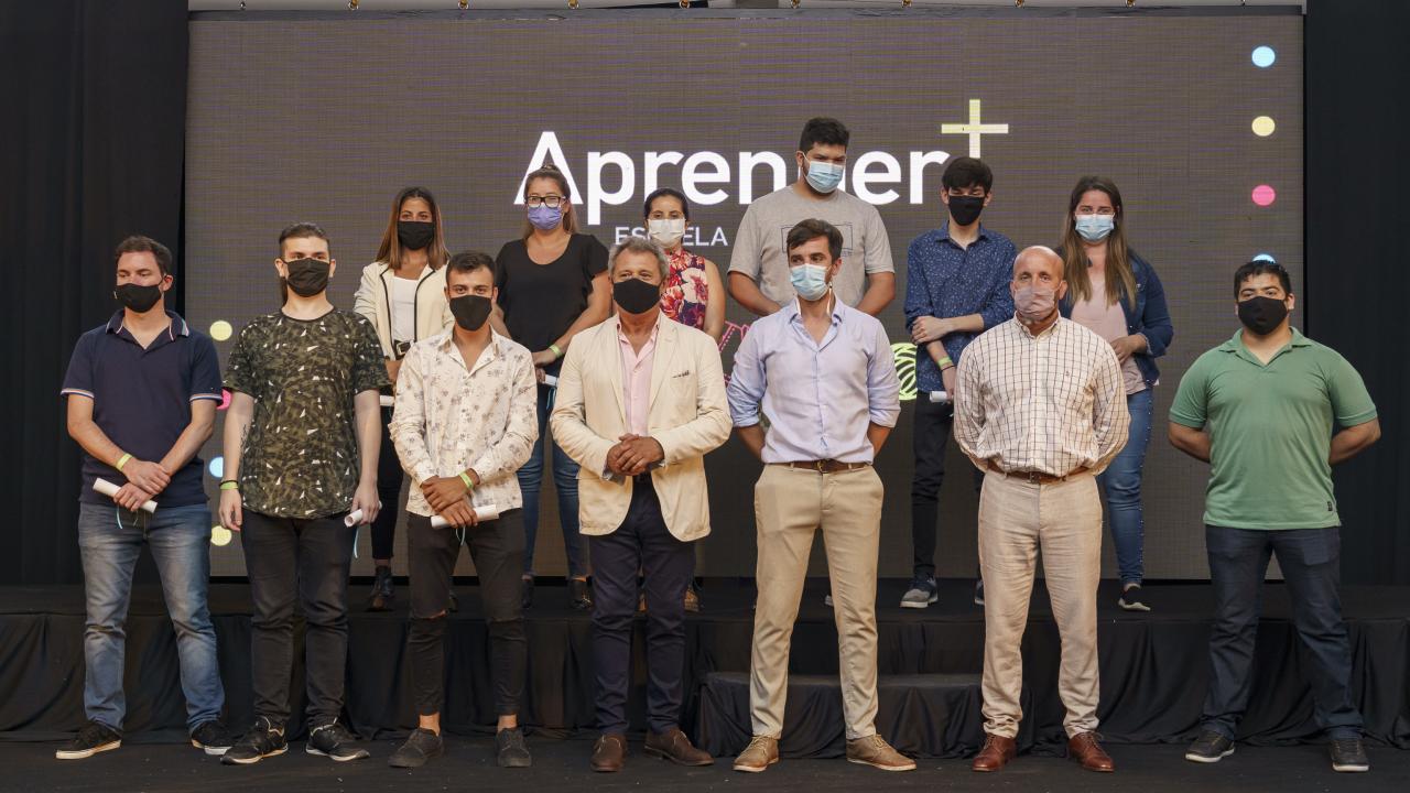 Grupo de personas con máscaras de pie en el escenario frente a una pantalla grande que muestra "Aprender+" con varios puntos de colores.