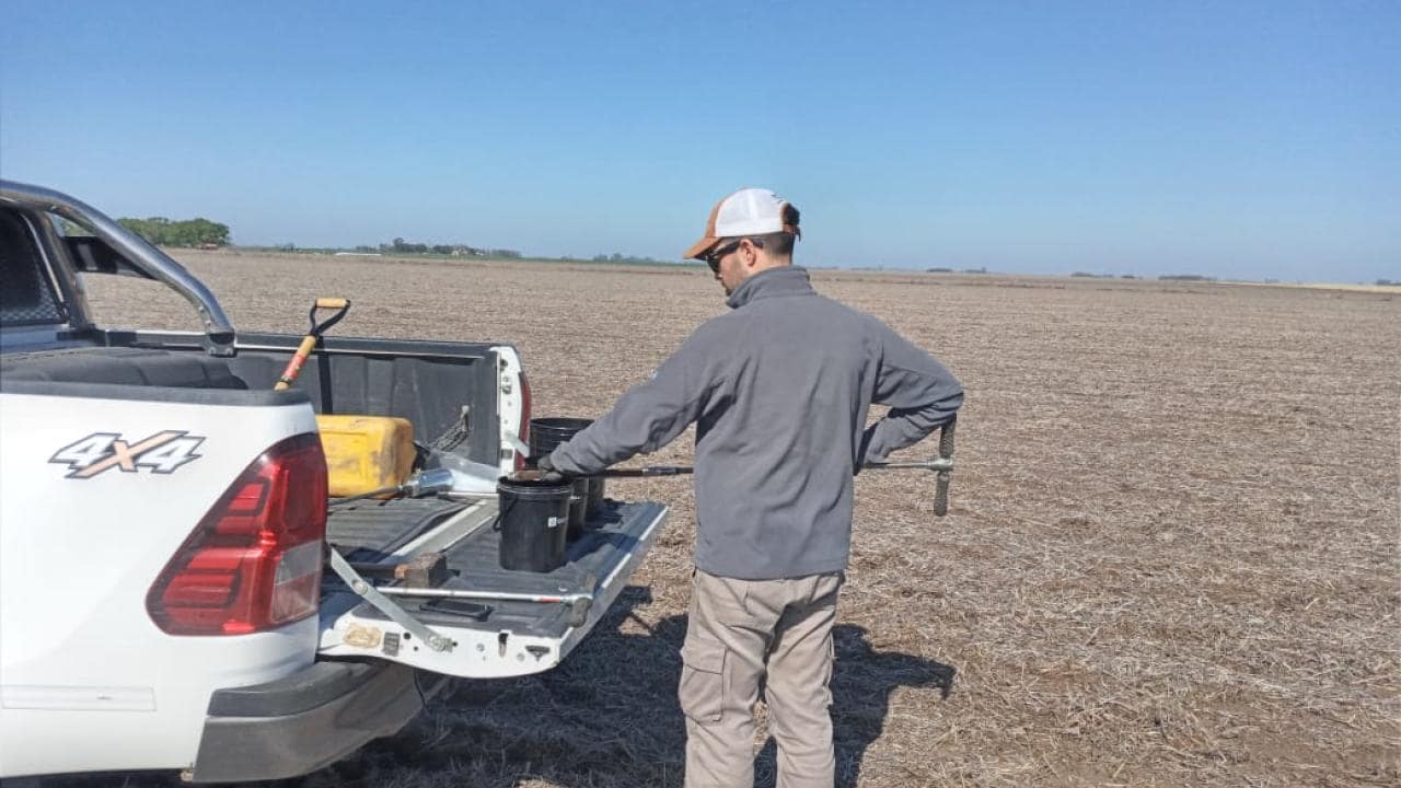 Una persona se encuentra junto a una camioneta en un campo arado, trabajando con equipos en la puerta trasera de la camioneta bajo un cielo azul claro.
