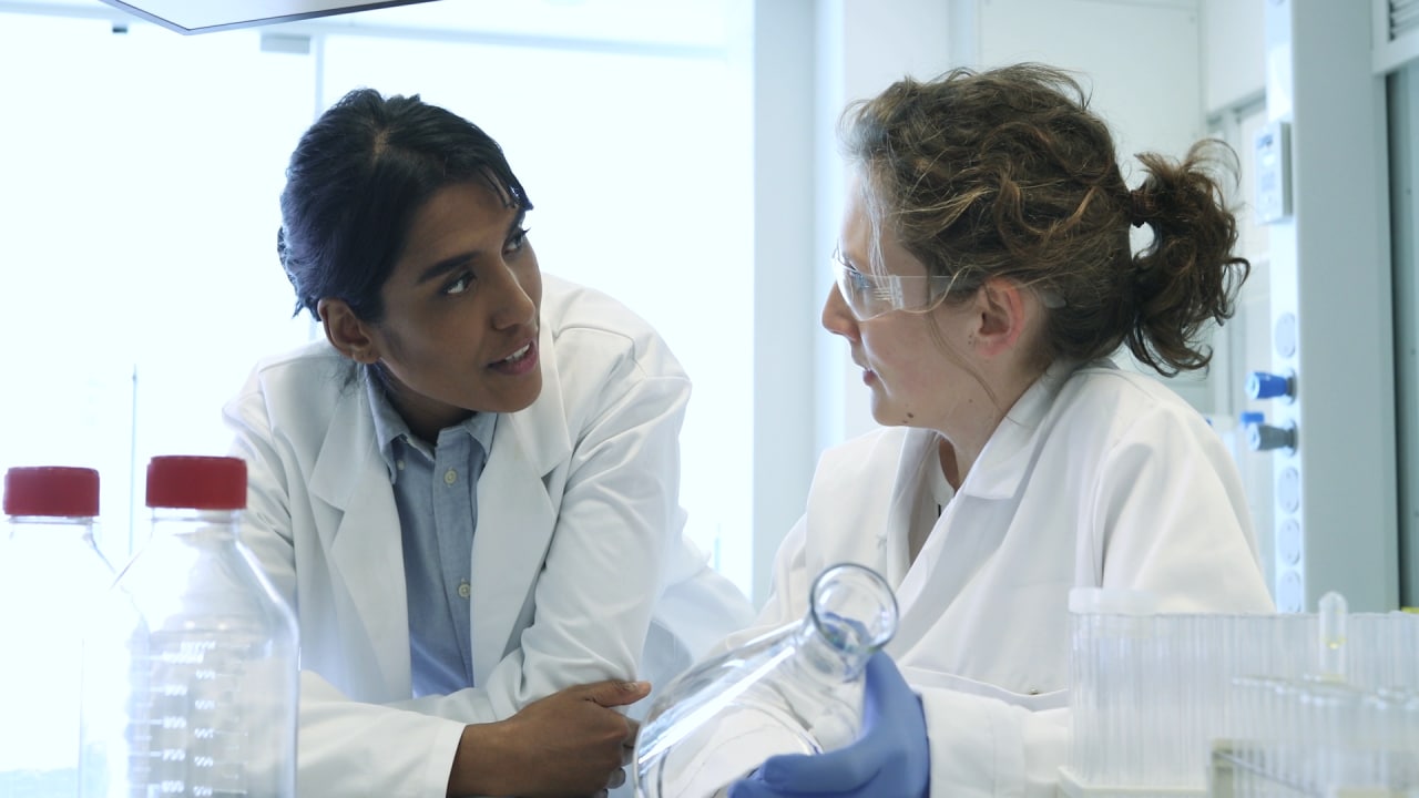 Dos científicos con batas blancas conversan en un laboratorio, rodeados de equipos y tubos de ensayo.