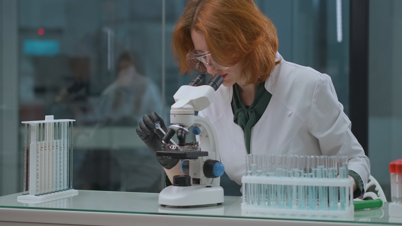 Una científica examina muestras bajo un microscopio en un laboratorio. Lleva gafas protectoras y guantes, rodeada de tubos de ensayo y equipos de laboratorio.