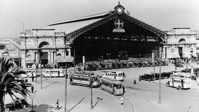 Estación de tren histórica con una arquitectura ornamentada, varios tranvías, automóviles y peatones en primer plano. Palmera en el lado izquierdo. Foto en blanco y negro.