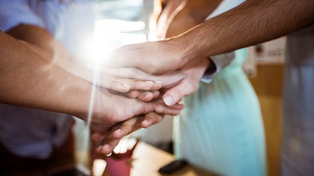 Varias manos apiladas juntas, simbolizando el trabajo en equipo, con la luz del sol brillando a través de ellas.