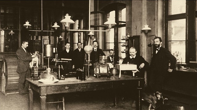 Un grupo de hombres en un laboratorio antiguo con equipo científico sobre la mesa, rodeado de grandes ventanales y luces colgantes.