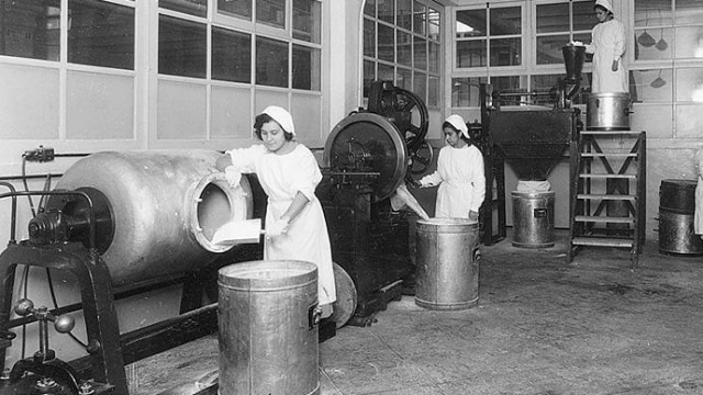 Las mujeres uniformadas operan equipos en una cocina industrial o una fábrica, trabajando con grandes contenedores y maquinaria.