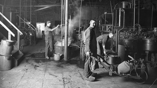 Los trabajadores operan maquinaria en un entorno industrial, del que se desprende vapor. Un trabajador se encuentra en primer plano sosteniendo una carretilla.