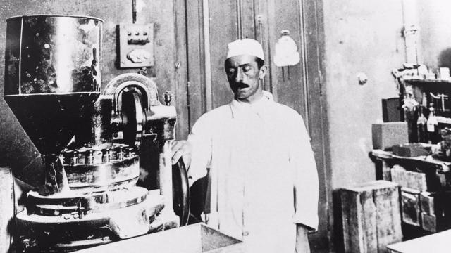 Un hombre vestido con ropa vintage se encuentra junto a una gran máquina industrial en un taller, rodeado de diversos equipos y herramientas.