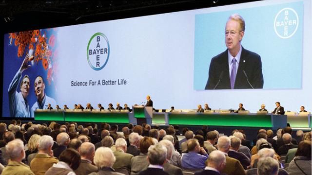 Un hombre habla en un podio sobre un escenario frente a un público. El fondo muestra el logotipo de Bayer, el eslogan "Ciencia para una vida mejor" e imágenes de investigaciones científicas.