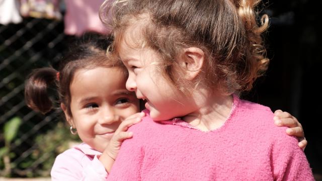 Dos chicas jóvenes, ambas con blusas rosas, sonríen y una abraza a la otra. Están paradas al aire libre con un fondo borroso de una cerca y ropa colgada.