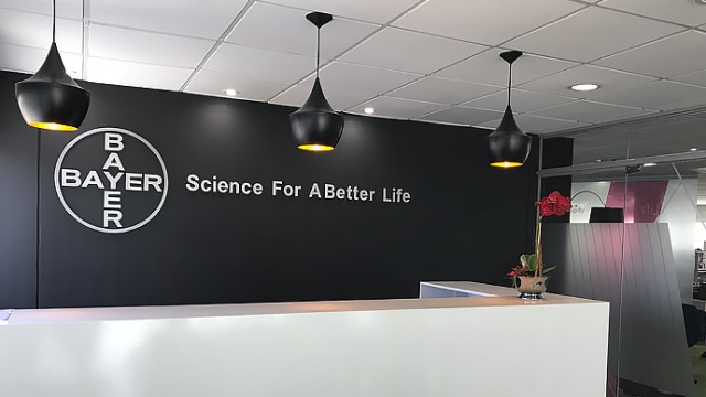Recepción de la oficina de Bayer con una pared negra con el logotipo de Bayer y el eslogan "Ciencia para una vida mejor". Tres lámparas colgantes negras cuelgan del techo y un arreglo floral sobre el escritorio.