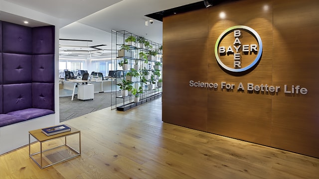Vestíbulo de oficina moderno con una pared de madera que muestra el logotipo y el eslogan de Bayer. A la izquierda hay un rincón para sentarse de color violeta y, al fondo, se ve el área de oficina de planta abierta con escritorios.