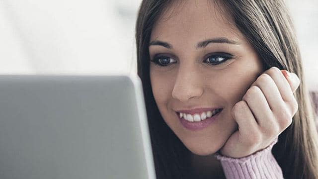 Una mujer con cabello largo sonríe mientras mira la pantalla de una computadora portátil con la mano apoyada en la mejilla.