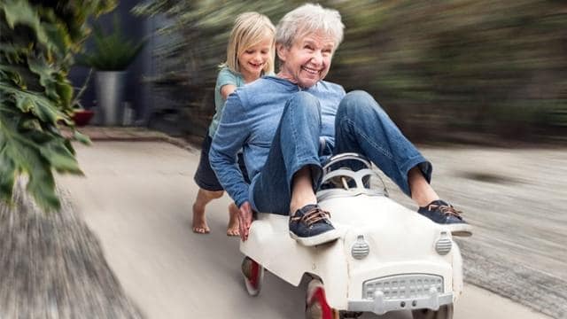 Un niño sonriente empuja a un anciano sentado en un pequeño coche de juguete; ambos parecen alegres mientras avanzan rápidamente por un camino.