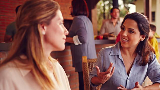 Dos mujeres conversan en una reunión, con otras personas de fondo. Una mujer hace gestos mientras habla y ambas parecen estar interesadas.