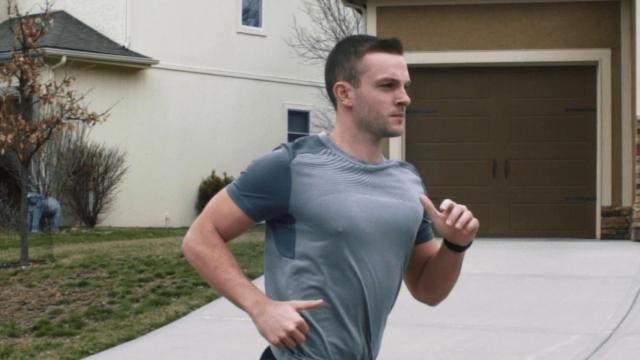 Persona haciendo jogging en una calle residencial, vistiendo una camiseta gris, con casas y un camino de entrada al fondo.