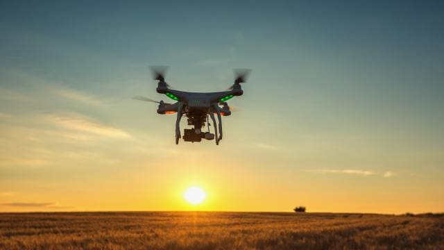 Un dron vuela sobre un campo al atardecer, con el sol iluminando el cielo y el paisaje.