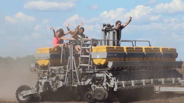 Tres personas saludando mientras están sentadas en una gran maquinaria agrícola en un campo bajo un cielo azul.