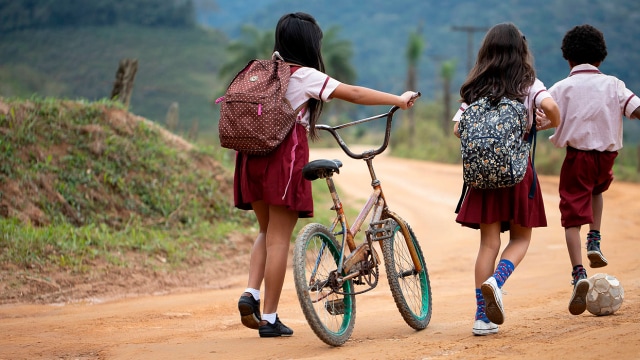 Tres niños con uniformes escolares caminan por un camino de tierra; dos llevan mochilas y uno empuja una bicicleta.