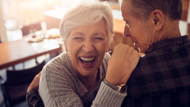 Una anciana ríe alegremente mientras abraza a un hombre en el interior, con una mesa y sillas de madera al fondo.