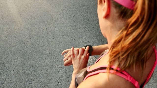 Una mujer vestida con ropa deportiva mira su reloj inteligente mientras está parada en una calle pavimentada.
