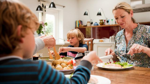Una familia de cuatro personas está comiendo en una mesa de madera en una cocina bien iluminada. Hay platos de comida frente a cada persona.