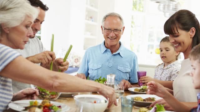 Una familia disfruta de una comida junta en una mesa de comedor, sonriendo y sirviendo comida. El ambiente es luminoso y alegre.