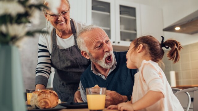 Un abuelo interactúa juguetonamente con su nieta en una mesa de comedor, mientras una abuela observa. Hay pan y jugo en la mesa.