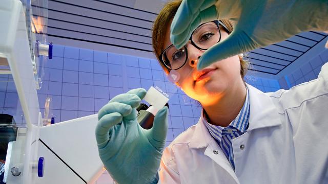 Un científico con bata y guantes examina un objeto pequeño, sosteniéndolo entre sus dedos, en un entorno de laboratorio.