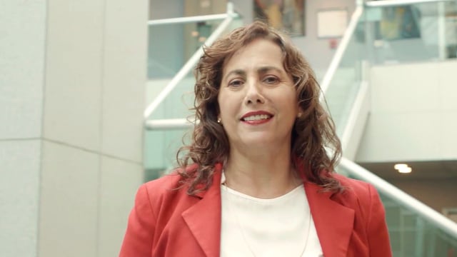 Una mujer con una chaqueta roja está de pie en el interior, sonriendo, con una escalera moderna y barandillas de cristal en el fondo.