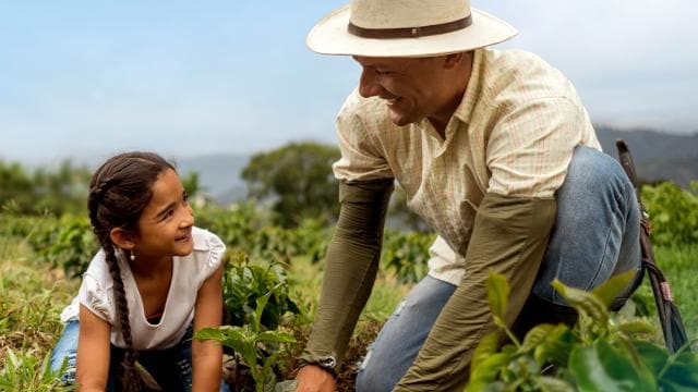 Un hombre y una niña están plantando en un campo. El hombre lleva un sombrero y la niña tiene trenzas. Ambos están sonriendo. En el fondo hay plantas verdes y un cielo nublado.