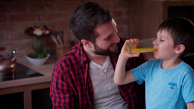 Un hombre con una camisa a cuadros sonríe a un niño que bebe jugo de naranja en una cocina.