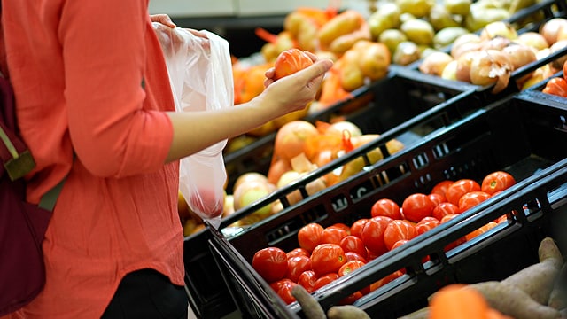 Una persona con una camisa naranja selecciona tomates de un contenedor de un supermercado, sosteniendo una bolsa de plástico.
