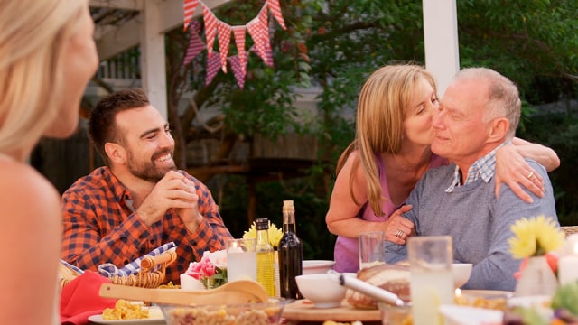 Grupo de personas disfrutando de una comida al aire libre; una mujer besa a un hombre mayor en la mejilla mientras otros sonríen.